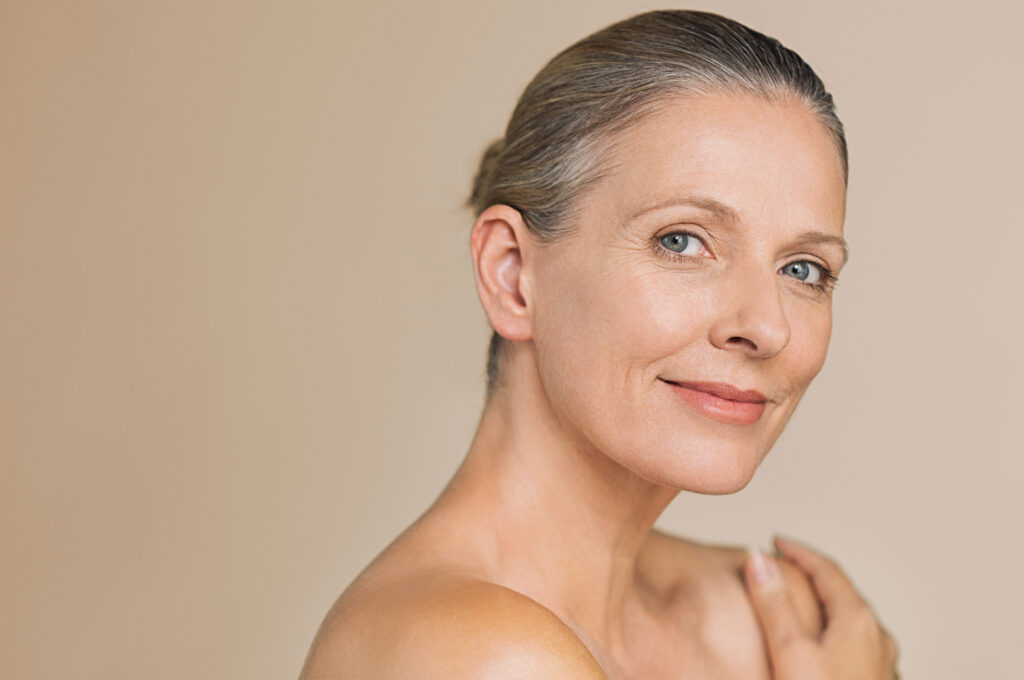 Smiling senior woman with bun hair and hand on naked shoulder. Portrait of beauty mature woman isolated over grey background with copy space looking at camera. Body and skin care concept.